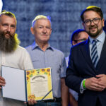 Na zdjęciu trzech mężczyzn. Jeden trzyma certyfikat z kolorowymi słonecznikami na żółtym tle. Tło jest ciemnoniebieskie z żółtym światłem.