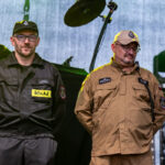 W sali bibliotecznej siedzą dzieci przy stole, przed nimi stoją słoiki w których robią dekoracje świąteczne. W tle stoi kobieta mówi do dzieci z nią regały z książkami