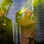 Przy drewnianym stole siedzą kobiety które robią dekoracje świąteczne z żywymi kwiatami w kolorze żółtym obol każdej leżą styropianowe jajka. W tle regały biblioteczne z książkami.