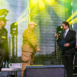 Na zdjęciu są cztery osoby na scenie. Tło jest ciemne z żółtym oświetleniem. Widać mundury i elegancki strój.