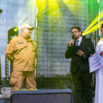 Na scenie cztery osoby: dwie w mundurach, jedna w garniturze z mikrofonem, kobieta w białej sukni trzyma certyfikat. Jasne reflektory na tle szarej kurtyny podkreślają postacie. Atmosfera oficjalna, oświetlenie w od