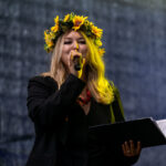 Osoba z wiankiem słoneczników na głowie śpiewa do mikrofonu. Ubrana na czarno, trzyma otwarty notes. Tło jest ciemne.