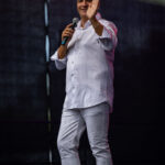 Osoba w białym stroju, z mikrofonem, macha na scenie. Ciemne tło sceny podkreśla sylwetkę. Światło pada z lewej strony.