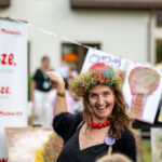 Na zdjęciu kobieta w kolorowym wianku, na tle banera Mazowsze. Ma na sobie czarną suknię i czerwony naszyjnik. W tle widoczne są dziecięce rysunki drzew.