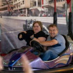 Na zdjęciu dwie osoby jadą w fioletowym samochodziku elektrycznym. W tle znajduje się dekoracja toru wyścigowego. Tło jest pełne kolorowych detali.