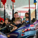 Na zdjęciu wesołe miasteczko, dzieci i dorośli w kolorowych samochodzikach na ciemnym tle. Kolorowe flagi nad nimi. W tle widoczna grafika z samochodem wyścigowym.