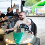 Na zdjęciu widać mężczyznę w okularach, jadącego zielonym samochodzikiem elektrycznym. Obok niego są inni bawiący się ludzie. Tło to wesołe miasteczko z zielonymi i białymi akcentami.