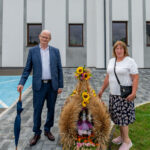 Starszy mężczyzna i kobieta stoją przy ozdobie z kłosów i słoneczników. Tło to biały budynek. Ozdoba jest kolorowa i ma tabliczkę z numerem trzy.