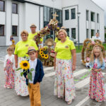 Grupa ludzi w kolorowych ubraniach przed nowoczesnym, białym budynkiem. Na pierwszym planie chłopiec z kwiatem słonecznika. Tło ozdobione wieńcem dożynkowym z kwiatów.