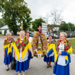 Grupa kobiet w kolorowych strojach ludowych stoi na tle drzew. Mają żółte chusty i niebieskie spódnice. Trzymają ozdobną koronę z kwiatów.