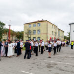 Na zdjęciu widać grupę ludzi w eleganckich strojach na placu, przed budynkami. Dominuje biel, czerń i czerwone akcenty. W tle drzewa i szare niebo.