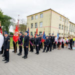 Na zdjęciu jest parada strażaków w mundurach, niosących czerwone sztandary. W tle widoczne jest szkolne, żółte budynek. Obok maszerują ludzie w eleganckich strojach.