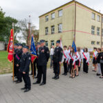 Grupa osób w uniformach i białych koszulach stoi przed budynkiem z flagami. Tło to żółty budynek i drzewa. Uroczysty charakter.