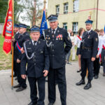 Grupa strażaków w czarnych mundurach stoi przed budynkiem. Tło: kremowy budynek, drzewa. Widoczne są kolorowe sztandary.