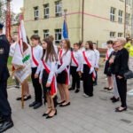 Grupa dzieci w białych koszulach i czerwonych szarfach stoi przed budynkiem szkoły. W tle są dorośli i inne osoby w kolorowych strojach. Dzieci trzymają sztandary szkolne, tworząc oficjalny nastrój.