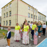 Grupa osób w kolorowych strojach i żółtych bluzkach z girlandami przed budynkiem. Chłopiec trzyma słonecznik. Tło to jasne, wielopiętrowe budynki.