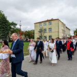 Na zdjęciu jest grupa ludzi w eleganckich strojach, niosących chleb i sól. W tle zielone drzewa i budynki. Niebo jest pochmurne.