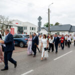 Parada ludzi w eleganckich strojach idących ulicą przy budynku. Mężczyzna niesie chleb na białej serwetce. Tło z samochodami, drzewami i budynkami.