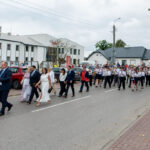 Grupa ludzi w eleganckich strojach i orkiestra maszerują ulicą. Budynki w tle są białe. Pogoda jest pochmurna.