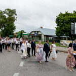 Ludzie idą w procesji po ulicy, w towarzystwie orkiestry dętej. Ubrani elegancko, jasne i ciemne kolory. W tle budynek, drzewa.