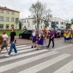 Na zdjęciu widać grupę osób w kolorowych strojach ludowych, przechodzącą przez przejście dla pieszych. Tło stanowią budynki mieszkalne i zaparkowane samochody. Widać też tradycyjne wieńce z kwiatów i zbóż.