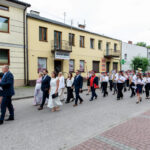 Parada na ulicy: ludzie w eleganckich strojach, orkiestra w białych koszulach i czerwonych mundurach. Tło kamienic.