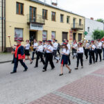 Orkiestra maszeruje ulicą w białych koszulach i granatowych mundurach, grając na instrumentach dętych. Tło stanowi żółty budynek z balkonami. Na chodniku widać czerwono-szarą kostkę brukową.