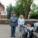 Na zdjęciu są trzy osoby: mężczyzna w mundurze, kobieta w jasnym garniturze i dziecko w wózku. W tle widoczne są drzewa i tablica informacyjna. Całość ma stonowane kolory, w tym dużo zieleni i brązu.