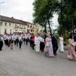 Na zdjęciu widać marsz ludzi w eleganckich strojach i orkiestrę. Uczestnicy niosą chleb na białych serwetach. Tło to budynki i drzewa, a niebo jest pochmurne.