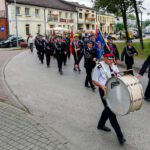 Parada strażaków w mundurach maszerujących wzdłuż ulicy z bębnem i kolorowymi sztandarami. Tło to małe budynki i zielone drzewa. Pogoda jest pochmurna.