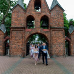 Ceglana brama z dzwonami, pod nią ludzie elegancko ubrani, otoczeni zielenią. Tło to drzewa i jasne niebo. Na brickowym bruku kroczą grupa.