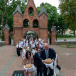 Grupa ludzi w eleganckich strojach niesie ozdobny chleb, idąc przez ceglaną bramę. W tle zielone drzewa i uczestnicy wydarzenia. Sceneria jest na zewnątrz, na wybrukowanym placu.
