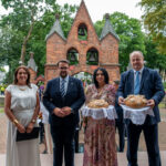 Cztery osoby stoją przed ceglanym budynkiem z dzwonami, dwie trzymają chleb na białych serwetkach. Tło zielone, drzewa.