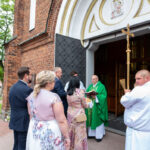 Grupa ludzi przed kościołem rozmawia z księdzem w zielonej szacie. Budynek z cegły. Widać dekoracje i drzewa.