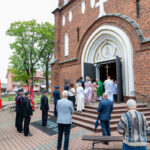 Kościół z czerwonej cegły, ludzie wchodzą przez drzwi, wokół drzewa. Grupa osób w strojach galowych trzyma czerwone flagi.