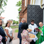 Grupa ludzi przy kościele, duchowny w zielonych szatach, obok ministrant. Kobieta z chlebem w tradycyjnym stroju. Tło: ceglany budynek.