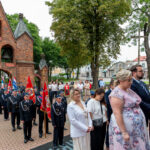 Grupa ludzi w eleganckich ubraniach stoi przed ceglaną bramą z dzwonami. Widać czerwone i niebieskie flagi. W tle drzewa oraz budynki na ulicy.
