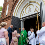 Grupa ludzi w strojach liturgicznych przed kościołem z cegły. Kapłan w zielonej szacie błogosławi uczestników. Tło zdobią drzwi z ornamentami i medalion z wizerunkiem Madonny.