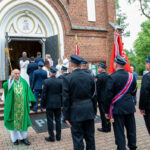 Grupa ludzi, w tym strażacy w mundurach, przed ceglaną ścianą kościoła. Ksiądz w zielonym ornacie kropi wodą. Flagi w tle.