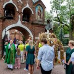 Na zdjęciu widoczny jest ksiądz w zielonym ornacie przed murowanym kościołem z dzwonami. Wokół niego znajdują się ludzie, niektórzy w odświętnych strojach, trzymający wieńce dożynkowe. Tło stanowią