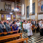 Grupa ludzi w kościele, ubrani formalnie, uczestniczą w ceremonii. W tle widać malowidła, okna i rusztowanie. Na podłodze wzory w szachownicę.