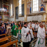 Procesja w kościele, ludzie w białych i zielonych szatach. Tło z freskami i żyrandolami. Uczestnicy stoją na czarno-białej posadzce.