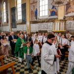 Odprawa kościelna z udziałem wiernych. Ludzie w kolorowych i białych strojach na tle fresków i witraży. Uroczysta atmosfera.