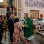 Na zdjęciu ksiądz w zielonym ornacie przyjmuje chleb od pary w kościele. Wnętrze jest ozdobione freskami i wzorami w ciepłych kolorach. W tle widoczne są świece i instrumenty dęte.