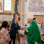Ceremonia w kościele, mężczyzna w garniturze przekazuje chleb księdzu w zielonej szacie. W tle malowidła i ozdoby. Obecni strażacy w mundurach.