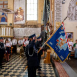 Grupa osób w mundurach strażackich w kościele trzyma niebieską flagę. Tło to kolorowe freski i witraże. Obok stoją dzieci w albach.