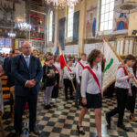 Grupa ludzi w kościele, młodzież z sztandarami, eleganckie ubrania. Wnętrze zdobione freskami i żyrandolami. Atmosfera uroczysta.