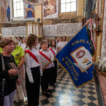 Grupa osób stoi w kościele, trzymając niebieski sztandar szkoły. Wnętrze zdobią freski i drewniane elementy. Tło jest jasne z dekoracjami.