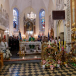 Wnętrze kościoła z uroczystością. Na pierwszym planie wieńce dożynkowe, w tle ołtarz z witrażami i malowidłami, dominują jasne kolory. Po prawej osoby w białych szatach liturgicznych.