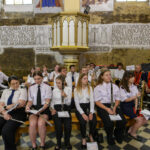 Grupa młodych muzyków w białych i ciemnych strojach siedzi w zabytkowym wnętrzu. Ściany ozdobione są freskami i napisami. Scena mieści się na tle kamiennej architektury.